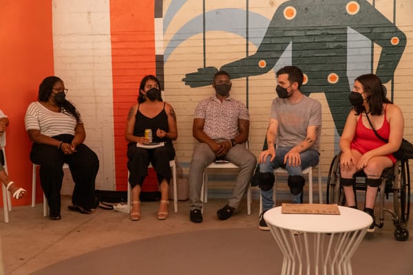 Five masked individuals seated in a semi-circle. The person on the right is sitting in a wheelchair, and Kelly wears leg braces. There's a colorful mural on the brick wall behind them.