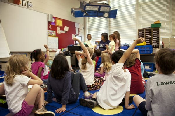 An audience of school children seated on the floor before a teacher at the front of the room. Some of the students are raising their hands.