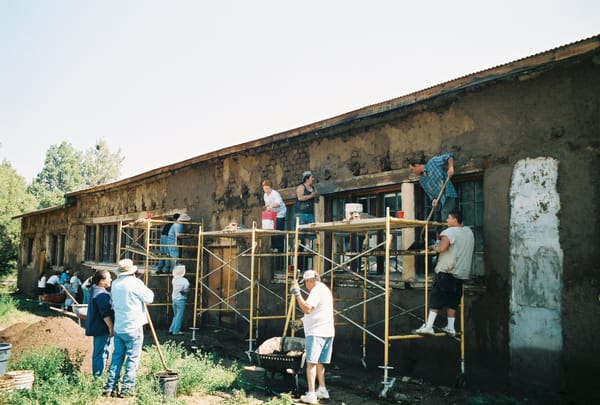 About a dozen people with scaffolding, shovels, wheelbarrows and other equipment, working on a long mud wall.