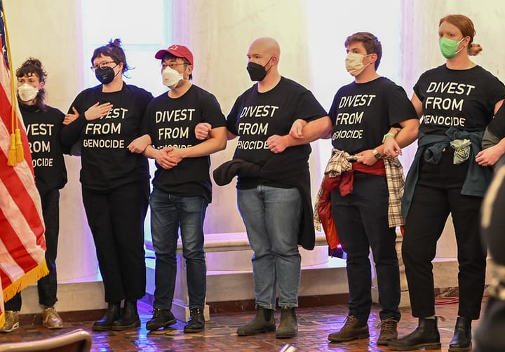 Six people wearing N95 or surgical masks and black tee shirts that read "Divest From Genocide" link arms facing the camera. An American flag is in the edge of the frame on the left.