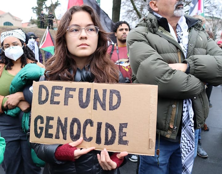 A demonstrator carries a "Defund Genocide" sign written in black marker on cardboard. Other demonstrators mill about.