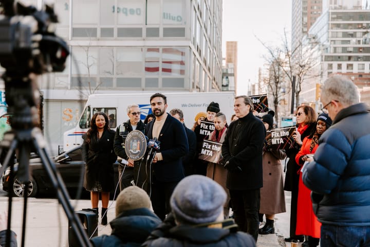 Zohran Mamdani stands and speaks at a small podium with a dozen people holding signs that read "Fix the MTA." News cameras and reporters face Mamdani in the photo's foreground.