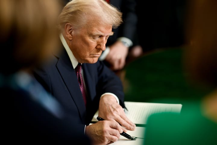 Donald Trump, wearing a dark suit and red tie, is seated, looking down at a desk, and signing a document. He is surrounded by blurred figures.