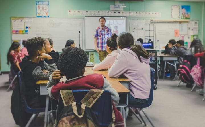 A classroom environment with about a dozen kids sitting at desks arranged in groups with a teacher standing at the front of the classroom. Many of the students are students of color.