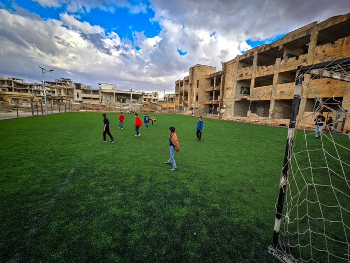 A group of children in brightly-colored clothes stand on artificial turf surrounded by damaged buildings. A slice of football net is visible at the edge of the frame.