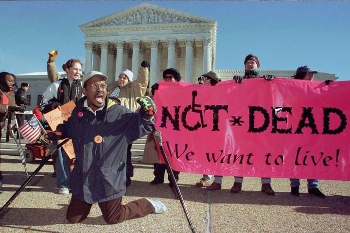A Black man holding crutches kneeling in front of the Supreme Court. A neon pink banner to the right reads, “NOT DEAD. We want to live!”