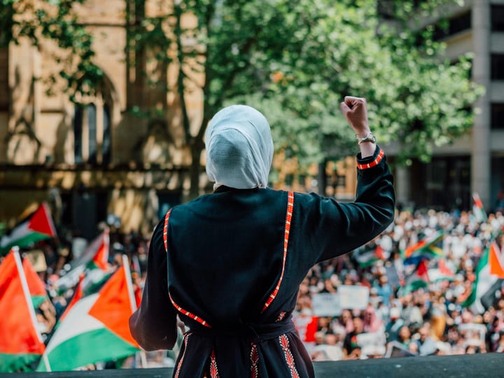 A person wearing a headscarf and dress with traditional Palestinian embroidery speaks to a crowd waving Palestine flags and placards.