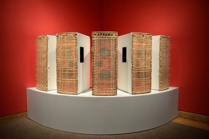 A collection of five red speakers with woven grass grates on their faces, arranged on a white pedestal in a corner against red walls.