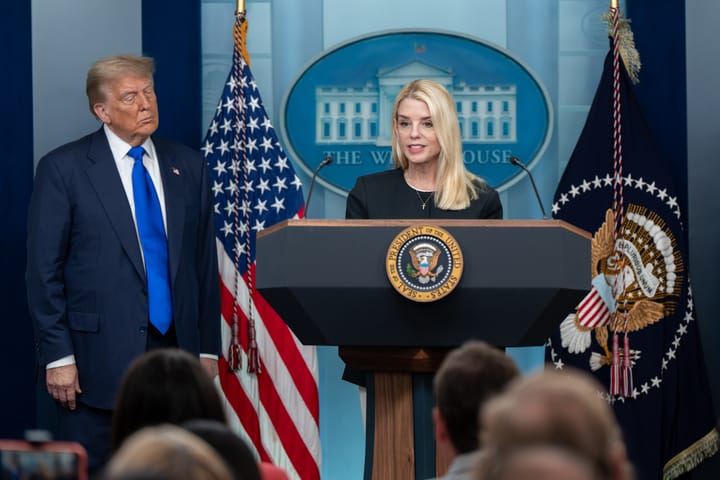 Pam Bondi, a blonde woman in black, stands at the podium in the White House Press Briefing Room. Trump, in a suit and blue tie, stands on the left side of the image, watching Bondi speak.