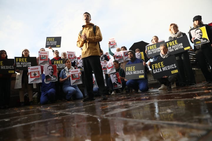 A group of people holding signs reading "Free Gaza Medics" and "Free Dr. Hussam Abu Safiya" gathered on wet paving stones. One person in a yellow rain jacket stands in the middle speaking.