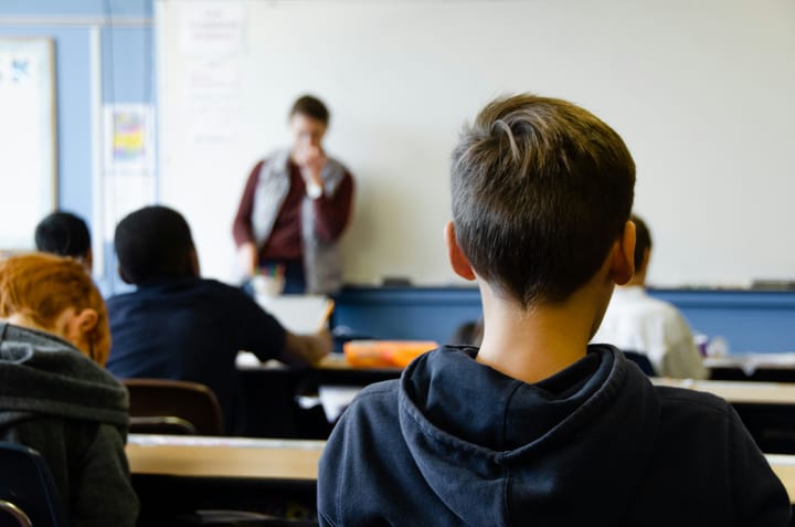 Students at desks with their backs to the camera; an adult stands at the front of the room before a whiteboard