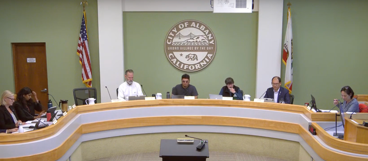 Seven people sitting at a long, curved table in front of a green wall with a City of Albany, California, crest hanging in the center. US and California flags hang on either side of the crest.