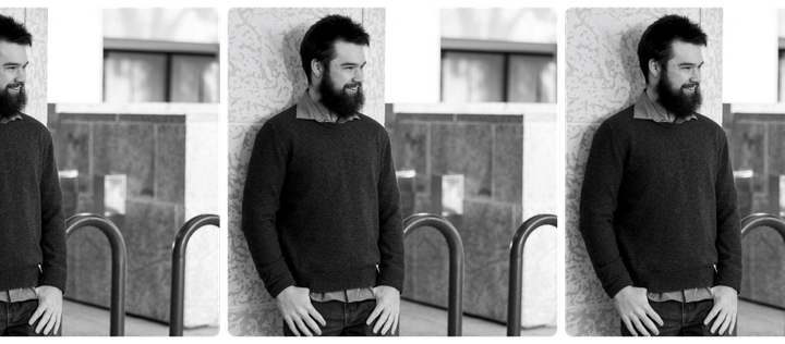 A black-and-white photo of John photographed from the waist up, wearing  jeans and a button-down shirt under a sweater. He's smiling and looking off camera.